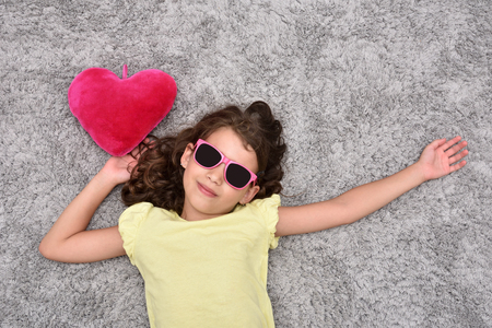 Smiling young girl with sunglasses and red plush heart lying on the carpet. Top view.の写真素材