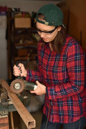 Woman with protective glasses using grinder on the metal pole in her own small workshop. Selective focus.の写真素材