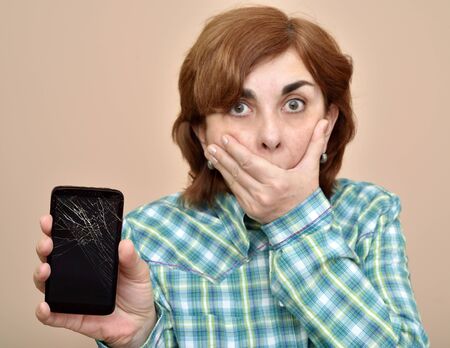 Surprised woman holding a broken smartphone in one hand and covering her mouth with the other hand. Focus on cracked display of the smartphone.の写真素材