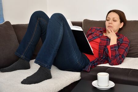 Beautiful woman wearing plaid shirt reading a book on the sofa in the living room at homeの写真素材