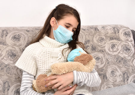 Sad and depressed young girl sitting on the sofa and holding her dog toy wearing medical protective face masksの写真素材
