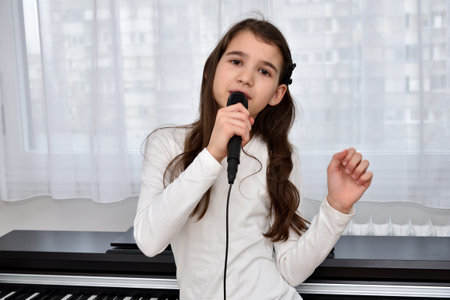 Cute young girl holding a microphone and singing at home in front of the window and electric pianoの写真素材