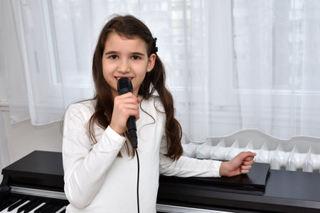 Cute young girl holding a microphone and singing at home in front of the window and electric pianoの写真素材