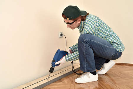Middle-aged woman with protective gloves and glasses using electric drill to make a hole in the...