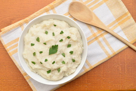 Mashed potatoes decorated with fresh chopped parsley in the white bowl, wooden spoon and dish cloth on the rustic wooden table. Close-up. Top view.の写真素材