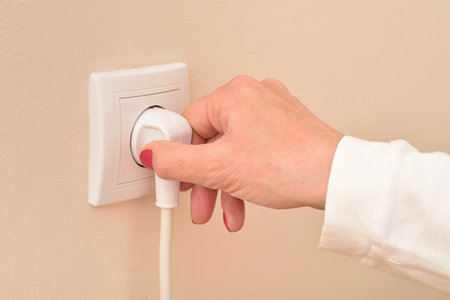 Hand of a woman holding and plugging or unplugging white electric cable in the white power socket on the pale orange wall. Copy space. Close-up. Concept of electricity consumption.の写真素材