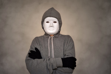 Unrecognizable person wearing hoodie, white mask and black gloves with crossed arms. Smoke or steam effect as a background.の写真素材