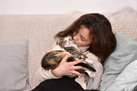 Young girl sitting on the sofa hugging and kissing her kitten. Friendship of child and animal. Keeping pets at home, care and love of animals concept.の写真素材