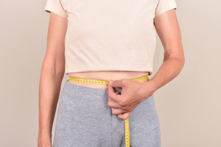 Slim mature woman measuring her waist with tape measure on the pale brown background. Body fit, dieting and weight loss concept.の写真素材