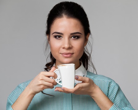 Frontal  portrait of an attractive brunette with a cup of coffee, looking straight, grey backgroundの写真素材