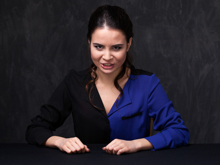 Portrait of an attractive brunette  looking straight. She is grimacing. Hands on the table. Grey backgroundの写真素材
