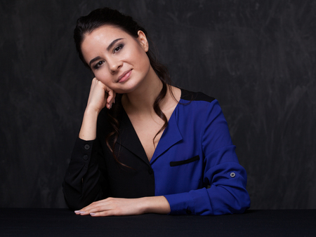 Portrait of an attractive brunette  looking straight and smiling. Her head is leaning on the fist. The other hand is on the table. Close up. Grey backgroundの写真素材
