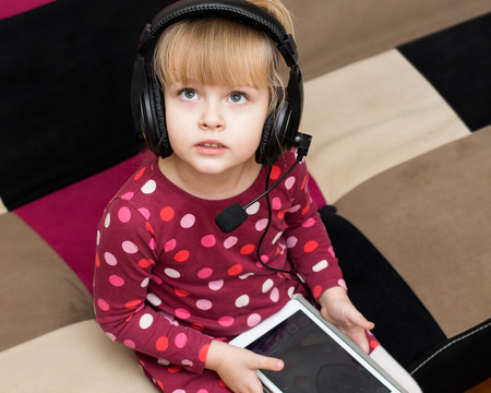 Little blond girl in the headphones with the tablet is looking up. Serious faceの写真素材