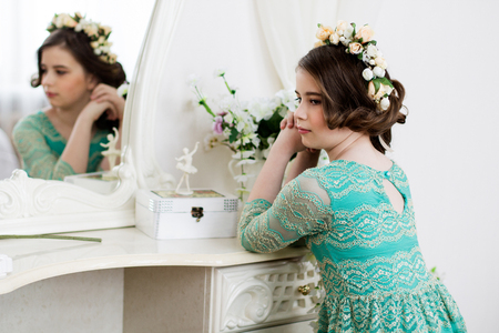 Little pensive girl in flower  wreath is sitting at the dressing table in front of a mirrorの写真素材