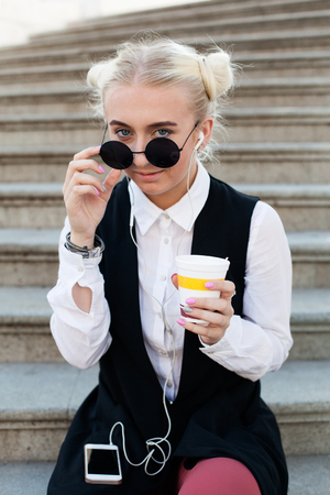 Young trendy woman in sunglasses listening to music and drinking  coffee on the stairsの写真素材