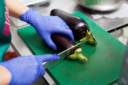 Woman's hands in gloves are cutting eggplants on the boardの写真素材