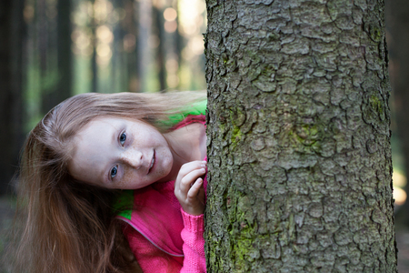 Beautiful red-haired girl with freckles looks from behind a tree in the forestの写真素材