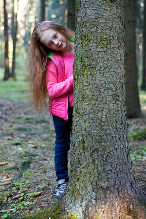 Little beautiful red-haired girl with freckles looks from behind a tree in the forestの写真素材