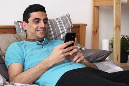 Happy arabic man using a smartphone, lying on bed at homeの写真素材