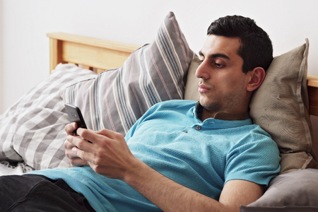 Young middle eastern male using a smartphone, lying on bed at homeの写真素材