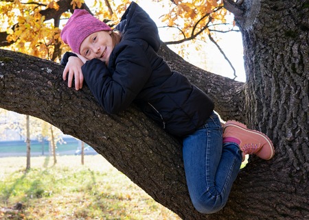 Happy little girl sitting on branch of big and smiling on sunny autumn dayの写真素材