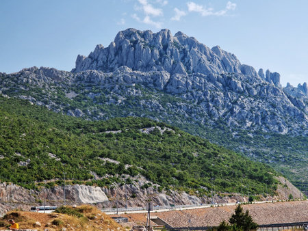 Rocky mountain with lush green forest and clear blue sky. A scenic summer landscape featuring rugged cliffs, vegetation, and a road in the foreground, ideal for travel or nature themes.の写真素材