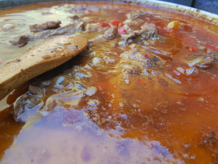 Close-up of bubbling beef stew with meat, vegetables, and rich broth in a pot. A wooden spoon rests in the liquid, showing the simmering cooking process. Rustic outdoor setting highlights traditional comfort food.の写真素材