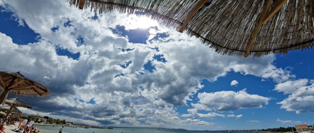 A vibrant beach scene with straw parasols under a dramatic sky filled with fluffy clouds. The sun peeks through, illuminating the coastline where people relax and enjoy the seaside on a warm summer day.の写真素材