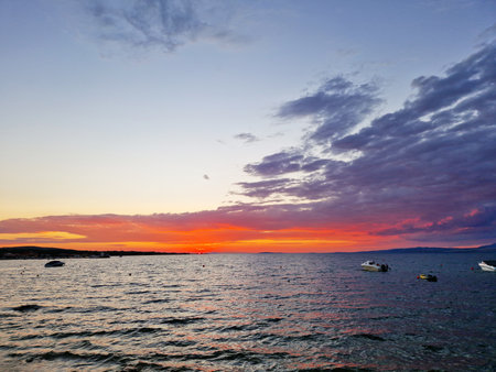 A stunning seaside sunset with vivid orange and purple hues reflecting on the water. Boats float calmly under a colorful sky filled with clouds, creating a peaceful coastal scene at dusk.の写真素材