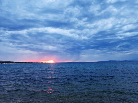 A dramatic seascape at sunset with dark stormy clouds over a calm ocean. The glowing horizon creates a striking contrast against the moody sky, reflecting warm hues on the water. A peaceful coastal scene in nature.の写真素材