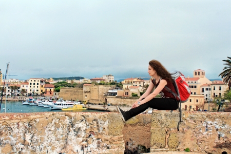 Young female tourist relaxing with her backpack with city of Alghero in the background, Sardinia,...