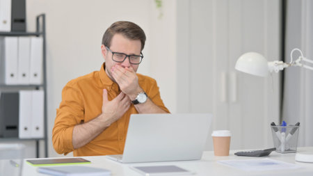 Young Man with Laptop Coughing in Officeの写真素材