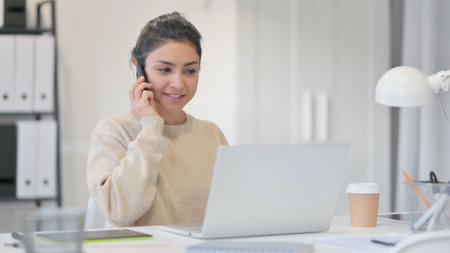 Young Indian Woman with Laptop Talking on Smartphoneの写真素材