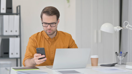 Young Man with Laptop using Smartphone in Officeの写真素材