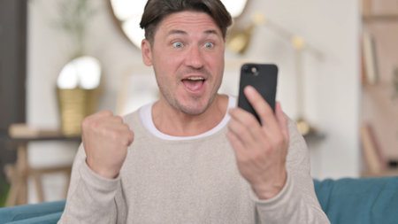 Portrait of Middle Aged Man Celebrating on Smartphone at Homeの写真素材