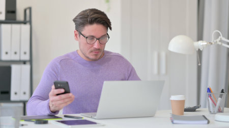 Middle Aged Man with Laptop using Smartphone in Officeの写真素材