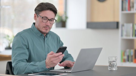 Middle Aged Man using Smartphone while Working on Laptopの写真素材