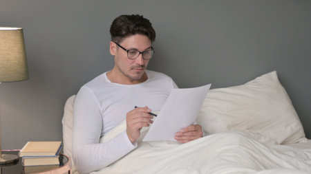 Hardworking Middle Aged Man Reading Documents in Bedの写真素材
