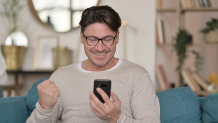 Portrait of Middle Aged Man Celebrating on Smartphone at Homeの写真素材