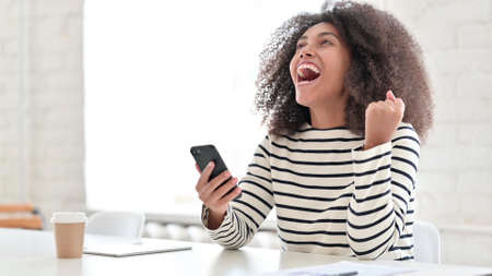Excited African Woman Celebrating Success on Smartphoneの写真素材