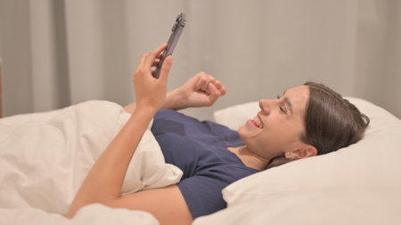 Side View of Young Latina Woman Celebrating Success on Smartphone in Bedの写真素材