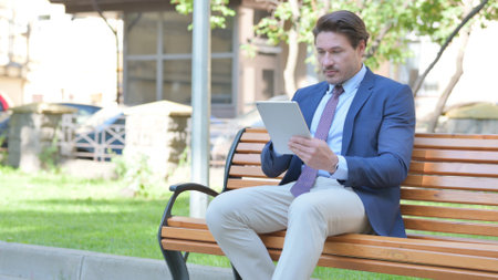 Middle Aged Businessman using Tablet while Sitting Outdoor on a Benchの写真素材