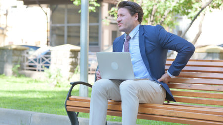 Middle Aged Businessman Using Laptop with Back Pain while Sitting Outdoorの写真素材