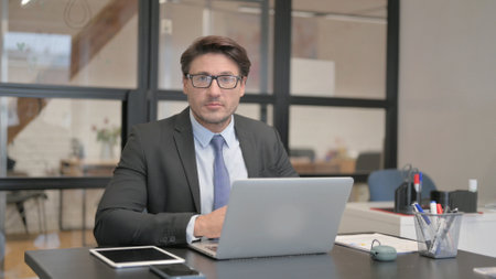 Businessman Looking at Camera while Working on Laptop in Office