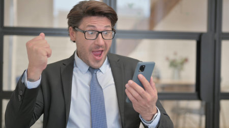 Businessman Celebrating Online Success on Phone while Sitting in Officeの写真素材