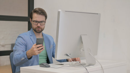 Young Man Using Smartphone for Working on Desktop