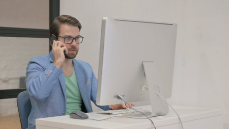 Young Man Talking on Phone while Working on Desktopの写真素材