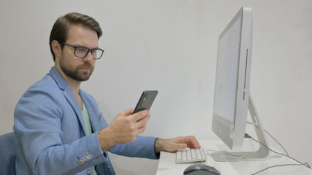 Middle Aged Man Using Smartphone for Working on Desktopの写真素材