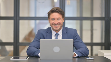 Middle Aged Businessman Smiling at Camera while Working on Laptopの写真素材