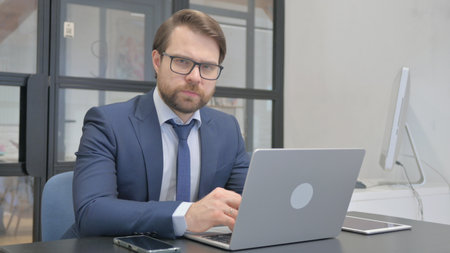 Young Businessman Looking at Camera while Working on Laptopの写真素材